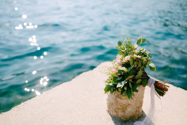 Bridal bouquet of pink protea, branches of eucalyptus tree, pink roses, capsella and white ribbons on the sea pier near the water — Stock Photo, Image