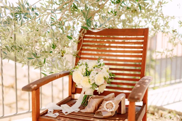 Ramo nupcial de rosas blancas, freesia, ramas de boj y cintas blancas cerca de los zapatos de la novia en la silla de madera debajo del olivo — Foto de Stock