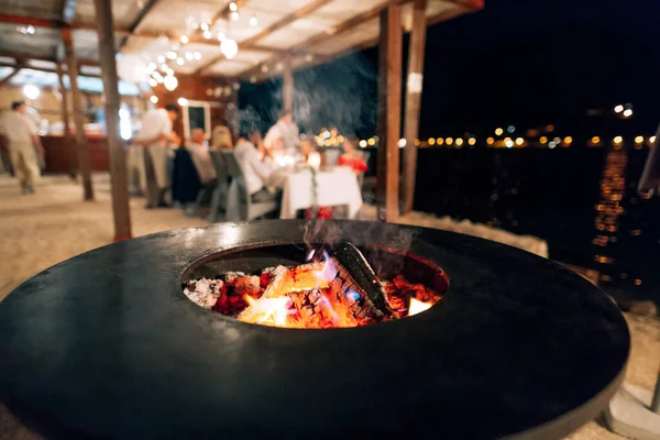 Grill plat noir, forme ronde, avec un trou avec un feu au centre. Dans le fond des gens qui dînent à la table. — Photo