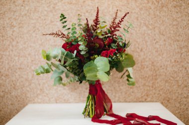 Burgundy şakayık buketi, kırmızı güller, okaliptüs ağacı dalları, eryngium, bebek nefesi, celosia ve kırmızı kurdele beyaz masada.