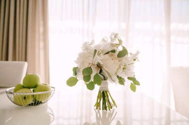 Beyaz şakayık demeti, tatlı bezelye ve okaliptüs ağacından dallar parlak beyaz bir masa üzerinde ve yanında yeşil elmalar olan bir kase.