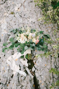 Gelin buketi beyaz ve kremalı güller, okaliptüs ağacı dalları, protea, eryngium, delphinium ve kayanın üzerinde duran beyaz kurdele.