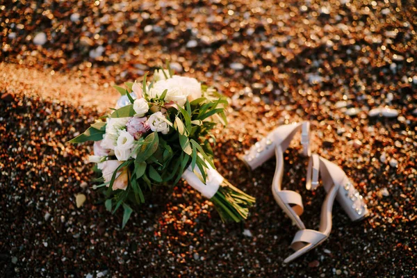Buquê de noivas de rosas brancas e rosa, peônias, ramos de cavalo de eucalipto, fitas brancas eustoma e os sapatos da noiva em uma praia de calhau — Fotografia de Stock