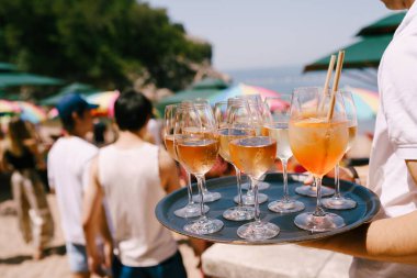 Elinde tepsi olan bir garson sahildeki bir partide kokteyl ve içki taşıyor. Büyük bir bardakta portakal kokteyli, yanında tüp Aperol spreyi ve bir bardak şampanya..