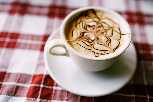 Primer plano del patrón de capuchino. Una taza de café con espuma en un platillo, sobre una mesa con un mantel rojo . — Foto de Stock