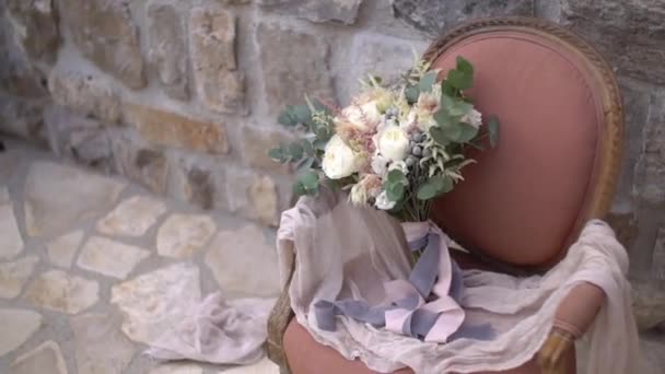 Bridal bouquet of white and cream roses, eucalyptus tree branches, pink and grey ribbons in the armchair the shoes of the bride near it — Stock Video