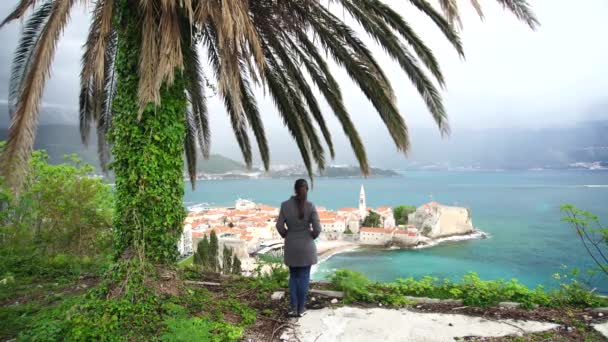 Dívka turista obdivuje staré město Budva v Černé Hoře z hory pod palmou. — Stock video
