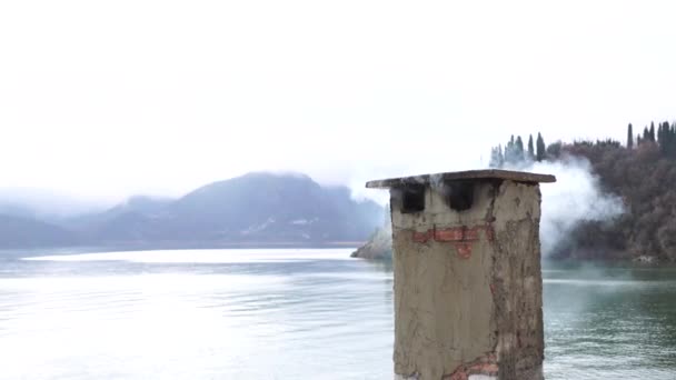 A brick stone chimney emits smoke against the background of the lake and mountains. — Stock Video