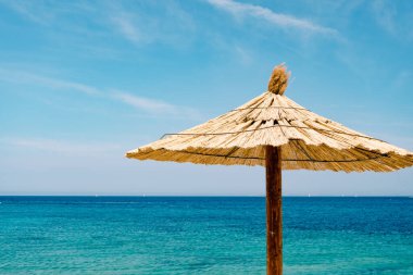 Hırvatistan 'ın Primosten kentindeki kumlu bir sahilde mavi gökyüzüne karşı sazdan bir şemsiye ve masmavi bir su..