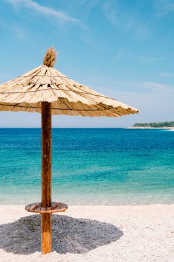 Hırvatistan 'ın Primosten kentindeki kumlu bir sahilde mavi gökyüzüne karşı sazdan bir şemsiye ve masmavi bir su..