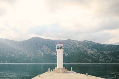 Karadağ 'ın Kotor Körfezi' ndeki Gospa od Skrpela adasında, Perast kasabası yakınlarındaki dağların arka planında, çerçevenin ortasında kırmızı bir fener bulunan beyaz taş deniz feneri..