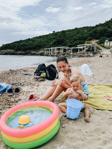 Mamá e hija en la playa. Juega en la arena con un cubo, cerca de una pequeña piscina inflable. — Foto de Stock