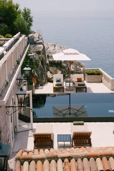 Budva, Montenegro - 12 juli 2020: Solstolar i trä under vita strandparasoller, nära poolen och kyrkan, med utsikt över havet. Sveti Stefan ön i Montenegro. — Stockfoto