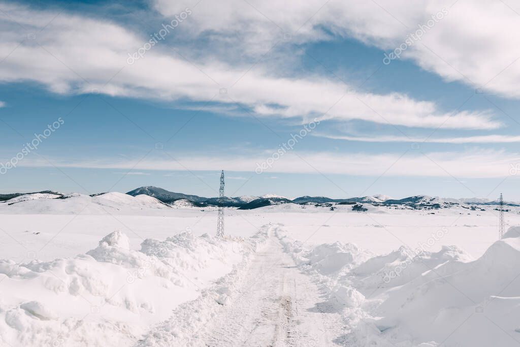 High power line poles on a snowy plain near a road with mountains on the horizon.