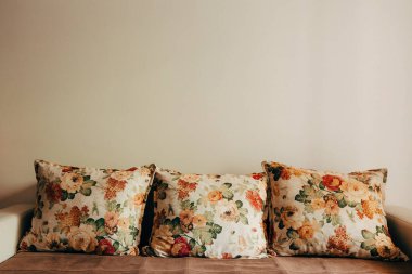 Kahverengi bir kanepede açık gri bir duvara karşı sıralanmış çiçeklerle işlenmiş desenli üç yastık..