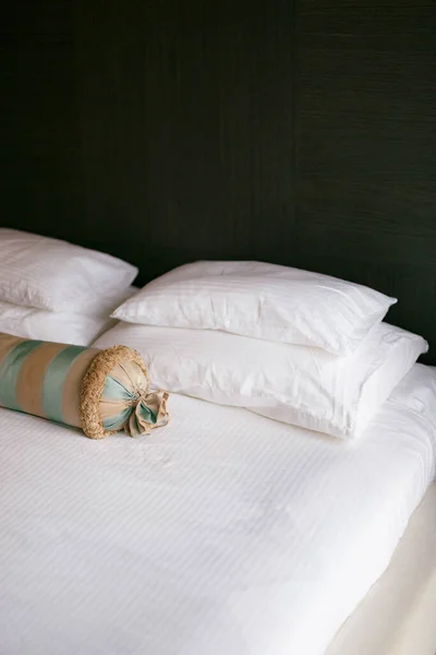 Una cama con una manta blanca y almohadas de diferentes tamaños y un cojín alargado en una habitación con paredes oscuras. —  Fotos de Stock
