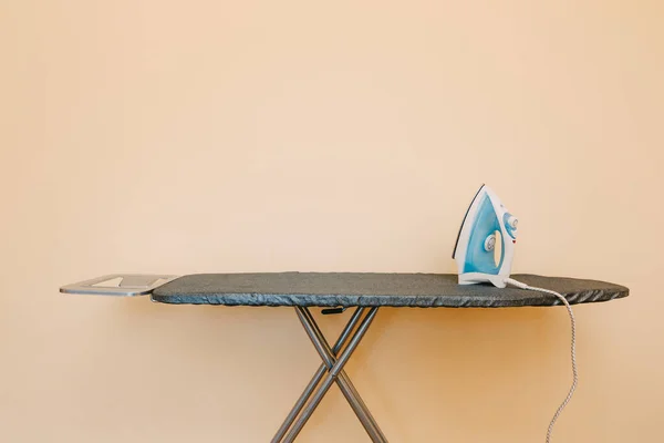 Una tabla de planchar con una plancha azul-blanca contra una pared beige. — Foto de Stock