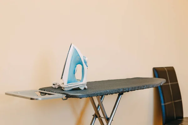 A blue iron on an ironing board with a cover near the chair in the room.