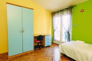 Bellagio, İtalya - 07 Haziran 2020: Sarı ve yeşil duvarlı bir genç için aydınlık oda, perdelerin arkasındaki balkona erişim, gardırop, masa ve yatak.