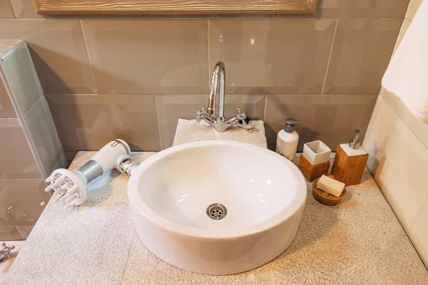 Close-up de um lavatório branco com sabão, produtos de higiene e secador de cabelo na bancada. — Fotografia de Stock