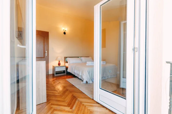 Una vista desde las puertas abiertas a una amplia habitación con una cama doble, lámparas de mesa y de pared, mesitas de noche con textura de pared clara. —  Fotos de Stock