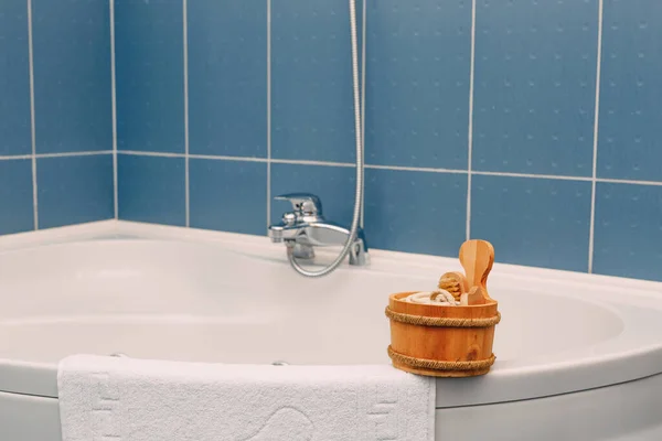 Деревянный ковш с щеткой, мочалкой и другими душевыми принадлежностями на краю угловой ванны возле махрового полотенца на фоне голубой плитки. — стоковое фото