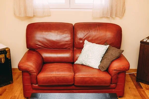 Раскладной диван из бордовой натуральной кожи с белыми и коричневыми подушками под окном с бежевыми шторами. — стоковое фото