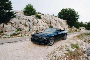 Tivat, Karadağ - 29 Temmuz 2020: Açık çatısı olan üstü açık siyah bir Ford Mustang, dağlarda, yolun kenarında, kayaların yanında.