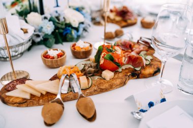 Doldurulmuş domates, jambonlu sosis ve peynirli tahta kesme masası kaseler, bardaklar, bir çiçek ve kaşık..