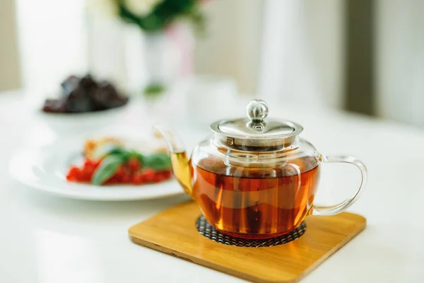 Крупный план маленького стеклянного чайника с чаем на деревянной доске под горячим блюдом на белом столе. — стоковое фото