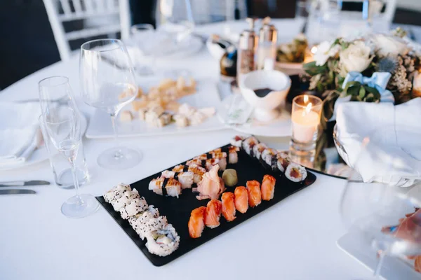 Primer plano de un plato negro para servir sushi y rollos sobre un mantel blanco sobre una mesa con vasos y platos sobre un fondo borroso. — Foto de Stock
