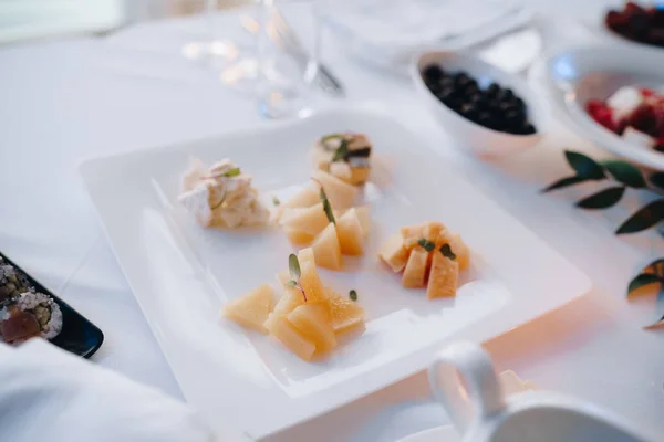 Un plato cuadrado con diferentes tipos de queso en rodajas sobre un mantel blanco junto a un tazón de aceitunas negras. — Foto de Stock