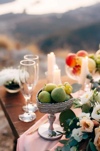 Sebuah mangkuk berwarna perak dengan buah ara dan anggur di atas meja dengan gelas sampanye, lilin dan buah-buahan dengan latar belakang kabur. — Stok Foto