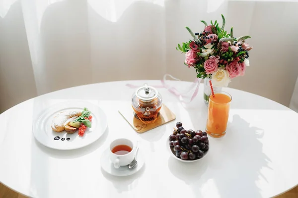 Uma mesa branca decorada com um buquê de flores junto à janela com uma cortina branca e um café da manhã de chá, croissant e uvas com anéis de ouro em um prato junto ao bule de chá. — Fotografia de Stock
