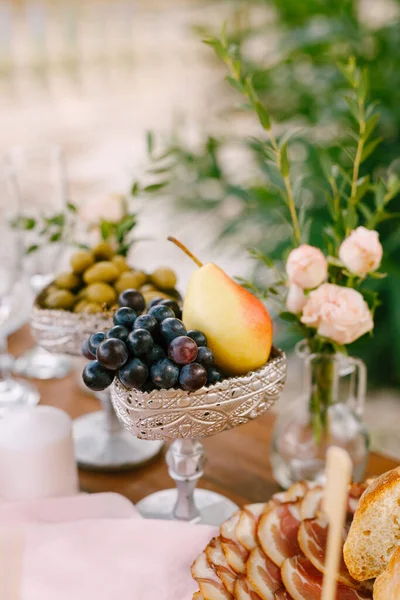 Un primer plano de un tazón de plata con uvas y una pera en la mesa con jamón, un jarrón con una flor y un tazón de aceitunas. — Foto de Stock
