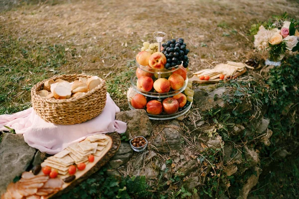 Whatnot with fruit, a basket with slices of bread and a board with cheese and meat cuts on stones in nature. — Stok Foto