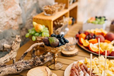 Tabaklarda dilimlenmiş jambon, peynir ve meyve bulunan bir masanın üzerinde altın bir kâse üzüm ve incir..