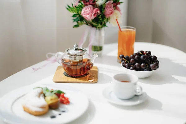 Zavrnik con té en un soporte de madera con anillos de boda en una mesa blanca con un plato, taza y tazón con uvas. — Foto de Stock