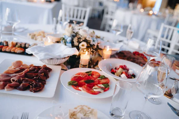 Platen met groentesalade, gesneden tomaten, prosciutto en sushi op een feestelijke tafel met een wit tafelkleed, glazen en kaarsen. — Stockfoto