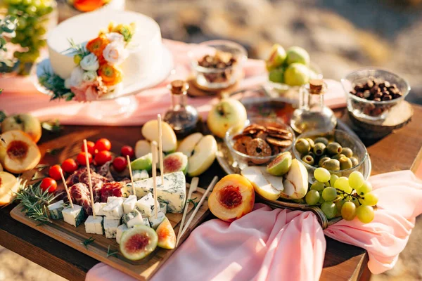 Papan potong dengan keju DorBlue, prosciutto dan tomat ceri, di atas meja kayu dengan buah-buahan dan kue pengantin putih dengan bunga. — Stok Foto