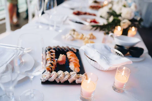 Placa negra para servir sushi y rollos sobre un mantel blanco sobre una mesa con vasos, toallas y velas encendidas. — Foto de Stock