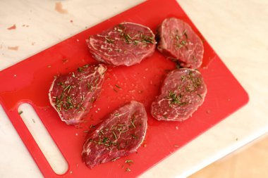 Kırmızı bir kesme tahtasına beş çiğ et parçası serpiştirilmiş biberiye ve kaba tuz..