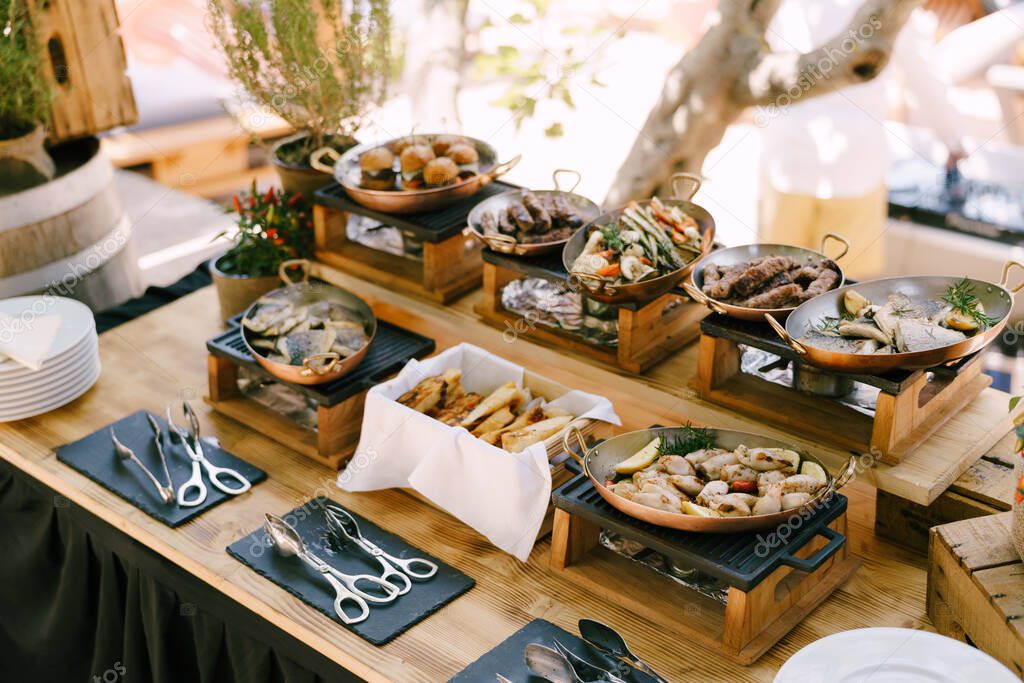 Ready meals in pans for the event on a wooden table with kitchen tongs scissors.