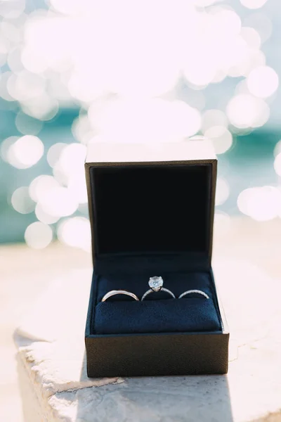 Goldene Eheringe und ein Verlobungsring mit einem großen Edelstein in einer schwarzen Samtschachtel auf verschwommenem Meeresgrund. — Stockfoto