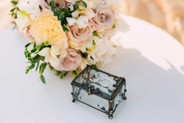 Gesloten doos met metalen smeden en gouden ringen voor de bruid en bruidegom op een witte tafel met een boeket van bruiloft bloemen. — Stockfoto