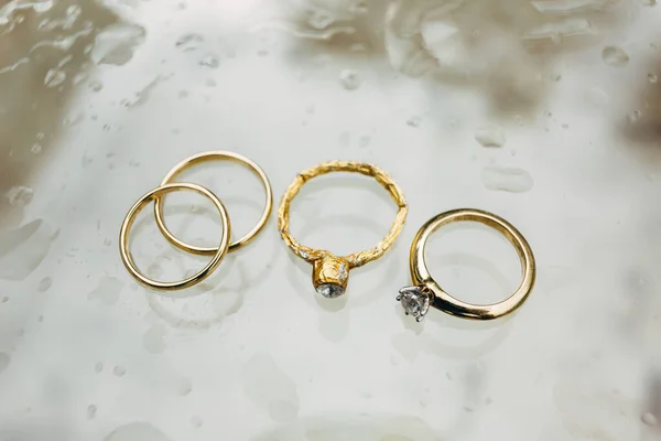 Close-up de anéis de casamento de ouro, um anel de noivado e um anel de folha metálica em um fundo cinza em gotas de chuva. — Fotografia de Stock