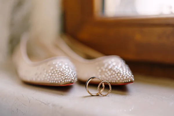 Detailní záběr snubních prstenů a baletních bytů nevěsty s korálky na rozmazaném pozadí na parapetu. — Stock fotografie