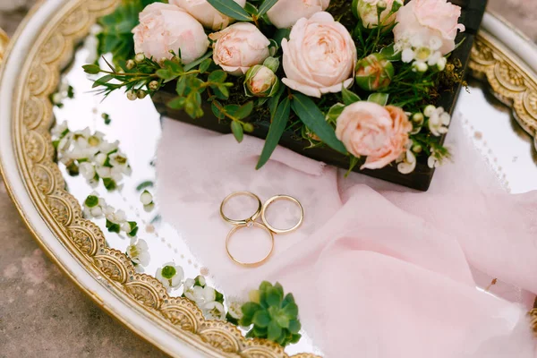 Due fedi nuziali per la sposa e lo sposo e l'anello di fidanzamento delle spose su un vassoio con motivi e un mazzo di rose rosa. — Foto Stock