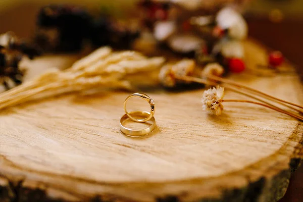 Close-up de anéis de casamento de ouro em um corte de madeira com espiguetas, flores secas e bagas em um fundo turvo. — Fotografia de Stock