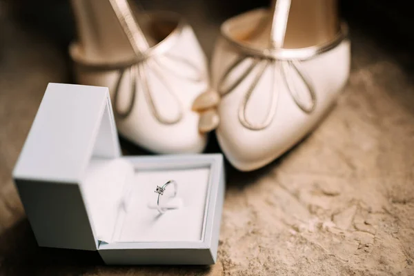 Δαχτυλίδι αρραβώνων σε ανοιχτό λευκό κουτί στο πάτωμα με γυναικεία νυφικά παπούτσια με πλεξούδες. — Φωτογραφία Αρχείου